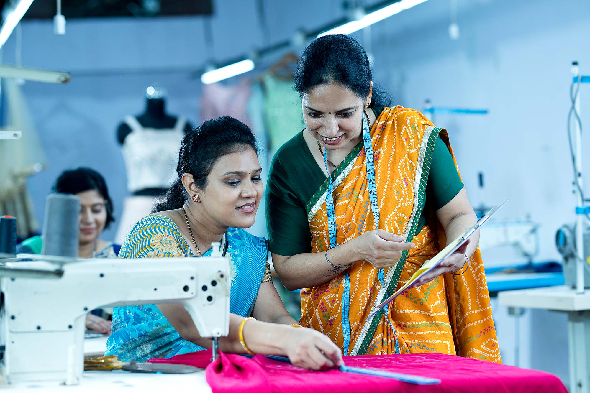 Inspector guiding female textile worker sewing garment on production line.